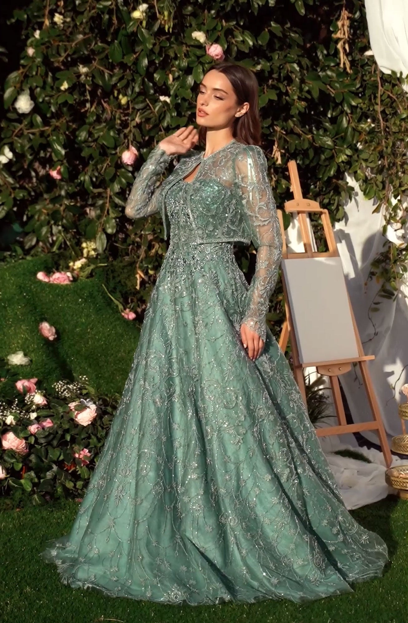 A woman in the Andrea & Leo MA104 Long Sleeve Long Formal Dress poses outdoors by an easel, surrounded by lush greenery and pink flowers.